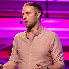 A man with short hair and some stubble wearing a pale shirt talking on stage in front of a brightly-coloured purple background.