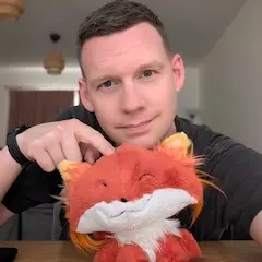 A short-haired man with a neutral expression on his face pointing down to a stuffed toy fox.