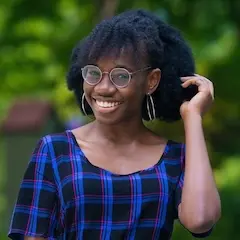 A smiling young woman with glasses wearing a plaid dress raising her hand to touch her long hair tied back.