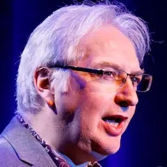 Profile of a middle-aged man with grey hair and glasses in mid speech.
