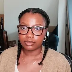 A young woman wearing glasses and her hair in braids sitting in a chair looking at the camera.