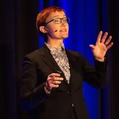 A woman with short red hair and glasses speaking animatedly on stage wearing a sharp suit with a floral shirt.